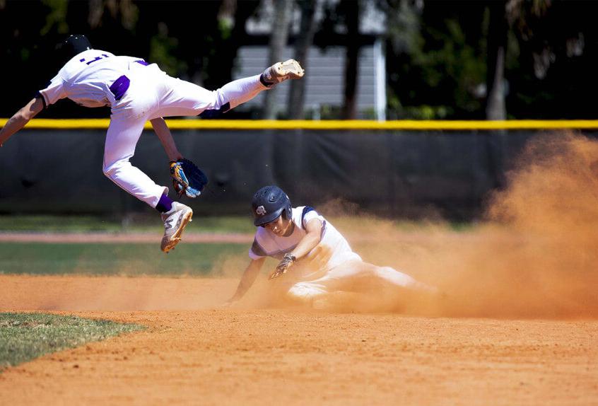 Poly Prep Baseball athlete sliding into base as the opposing team member leaps in the air for the ball